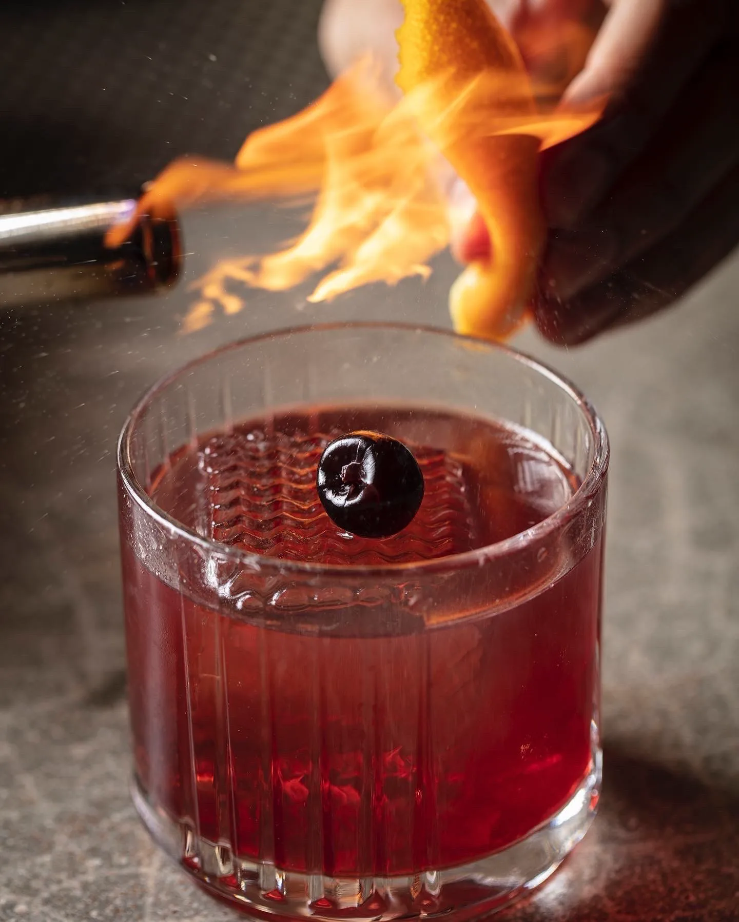 A deep-red craft cocktail with a cherry as a bartender flames an orange peel over it at Azabu